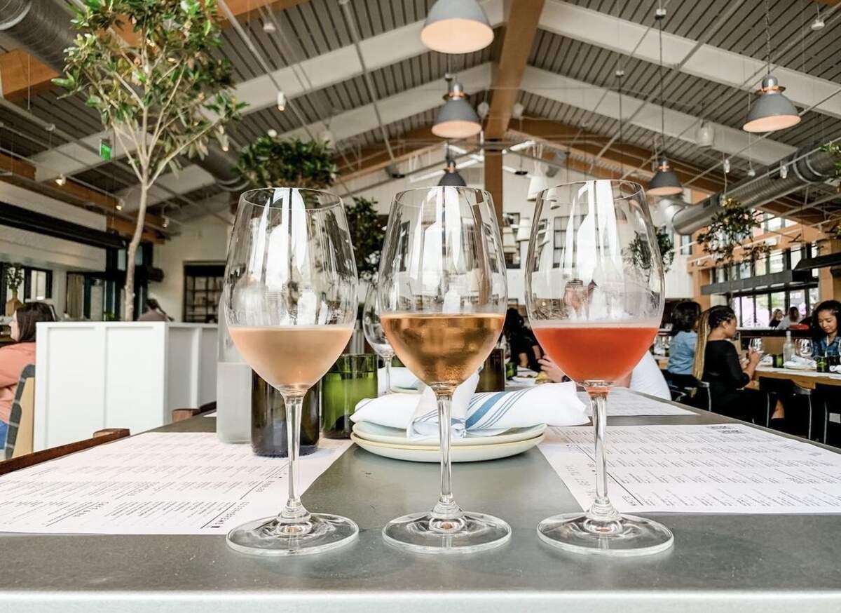 Three wine glasses filled with different shades of rosé wine, ranging from pale peach to deep pink, are lined up on a metal tabletop. In the background, the bright, airy interior of the restaurant features tall ceilings, indoor trees, and patrons dining at tables.