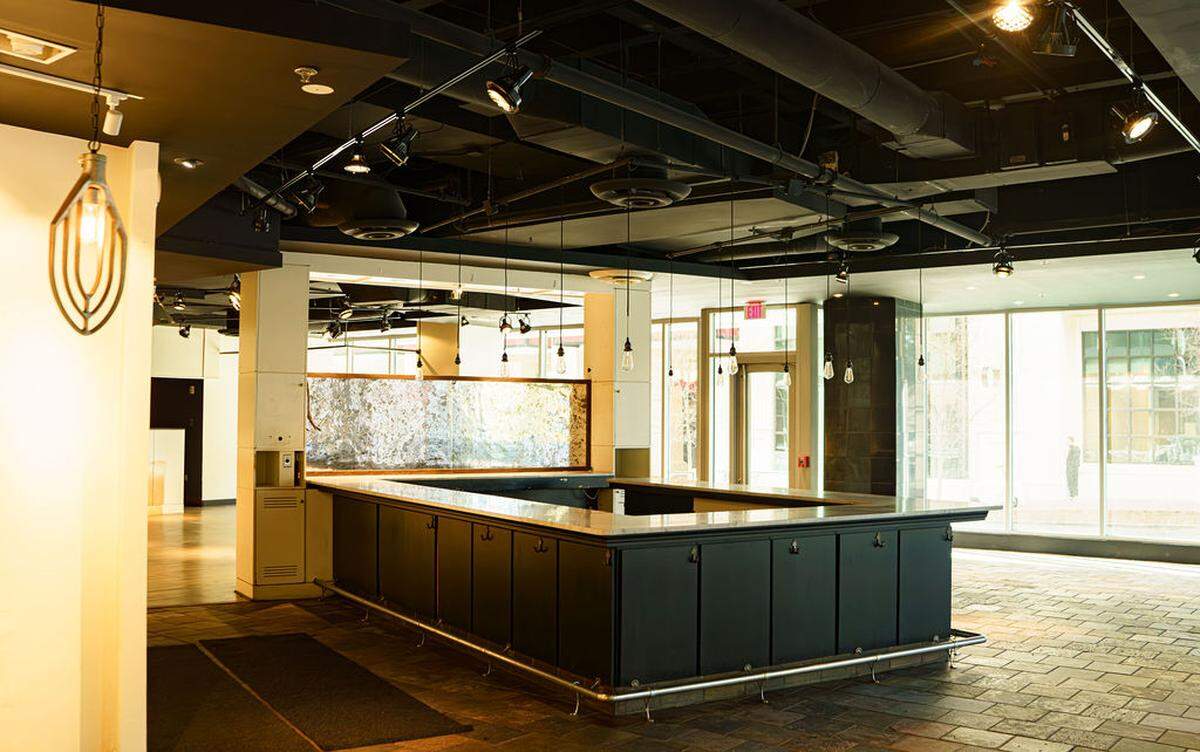 An empty, wide-angle view of a commercial kitchen space with an industrial stove and hood on the right. The left side features a long service window with stainless steel shelving and white walls showing multiple electrical outlets and signs of prior use or renovations.