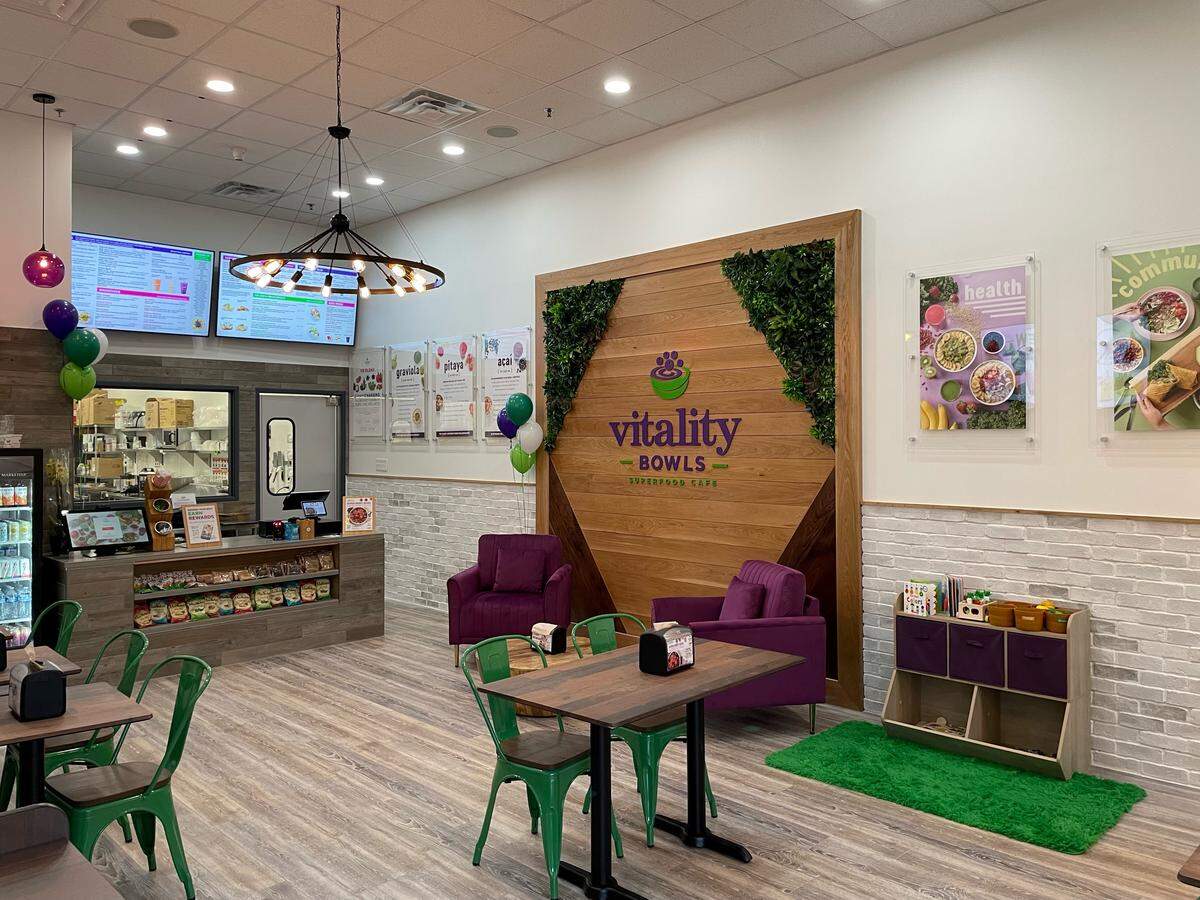 The interior of a Vitality Bowls café features a modern seating area with purple armchairs, green metal chairs, and a children’s play nook. A large wooden wall with a moss-lined logo serves as a focal point behind the dining tables.