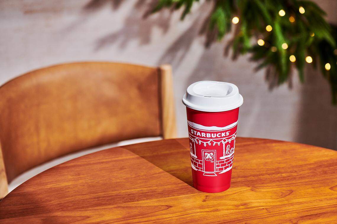 A reusable red Starbucks holiday cup with a white lid rests on a smooth, warm-toned wooden table in a cafe setting. In the background, a curved wooden chair, a light-colored wall, and a piece of greenery with small string lights are visible.