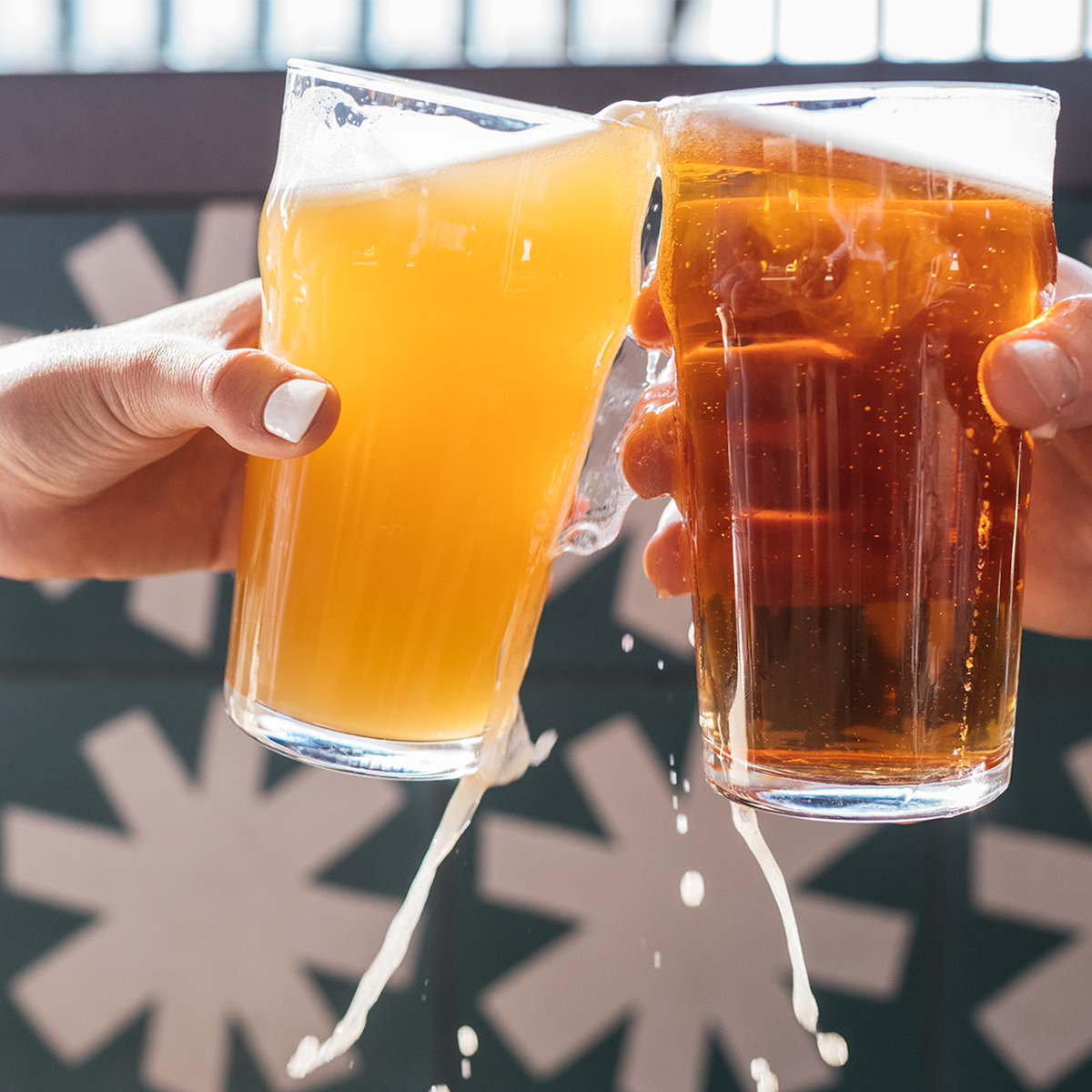 Two people clink glasses of beer in a celebratory toast, causing some of the liquid to splash over the sides. The background features a modern wall with white star-like patterns, highlighting a lively happy hour vibe.