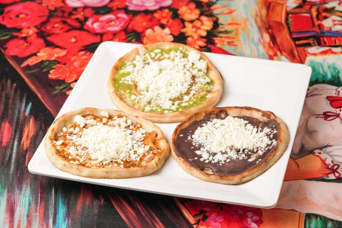 A white square plate holds three round, flat picaditas, each spread with a different base: green salsa, orange salsa, and dark bean paste. Every picadita is generously topped with a heap of crumbled white queso fresco and diced onions. The plate rests on a decorative surface featuring traditional Mexican-style illustrations and floral patterns.