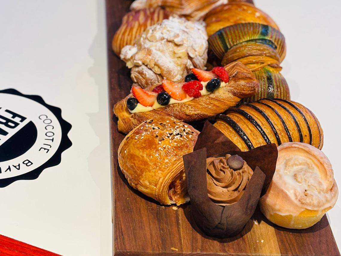 A variety of colorful and flaky French pastries, including croissants, pain au chocolat, and fruit tarts, are artfully arranged on a dark wooden serving board. A circular black-and-white “FRANCE BAKERY” logo is visible on the white table surface to the left.