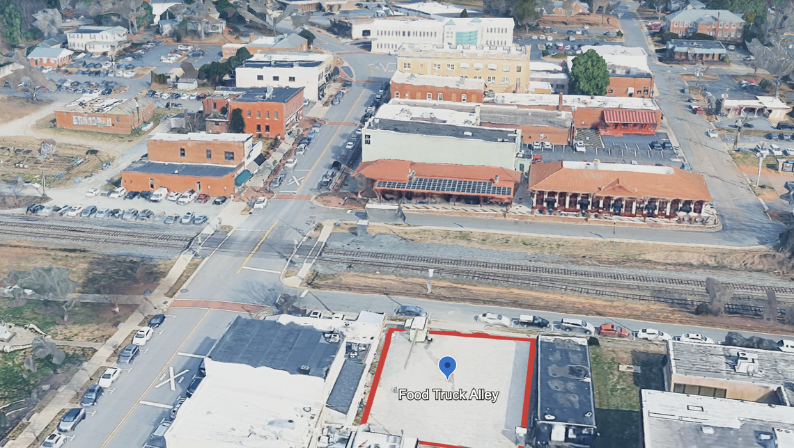This second angled aerial view shows the same small town’s downtown, but from a different perspective facing roughly south. The red outline and a blue location pin marking “Food Truck Alley” is visible in a lot across the street from a set of railroad tracks and several larger commercial buildings.
