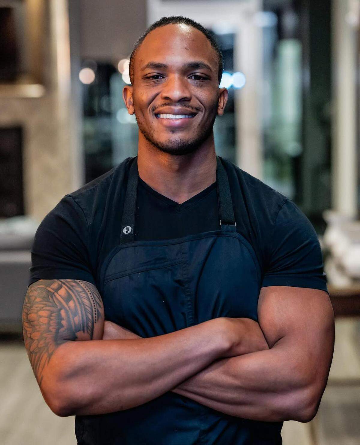 A smiling man with a large tattoo on his right arm stands confidently with his arms crossed while wearing a black t-shirt and apron. He is positioned in the center of the frame against a softly blurred, modern indoor background.