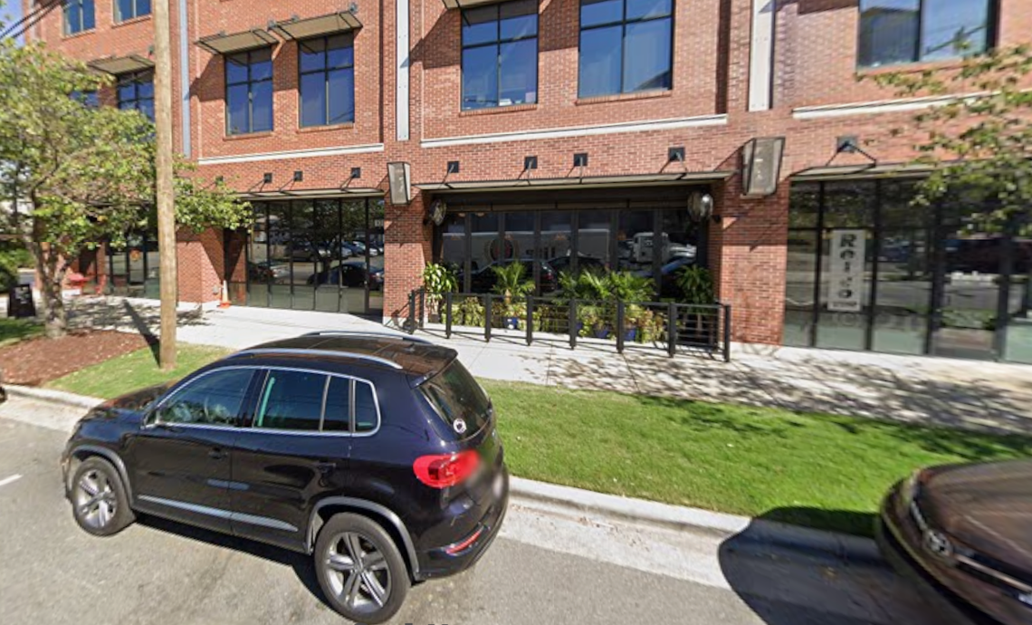 A street-level view of a modern, multi-story building with a red brick facade and large dark-framed windows. A dark-colored SUV is parked on the street in the foreground, with a grassy sidewalk strip separating it from the building. The ground level features a restaurant with large glass panels and a small fenced-in patio area with potted plants.
