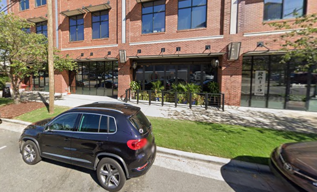 A street-level view of a modern, multi-story building with a red brick facade and large dark-framed windows. A dark-colored SUV is parked on the street in the foreground, with a grassy sidewalk strip separating it from the building. The ground level features a restaurant with large glass panels and a small fenced-in patio area with potted plants.
