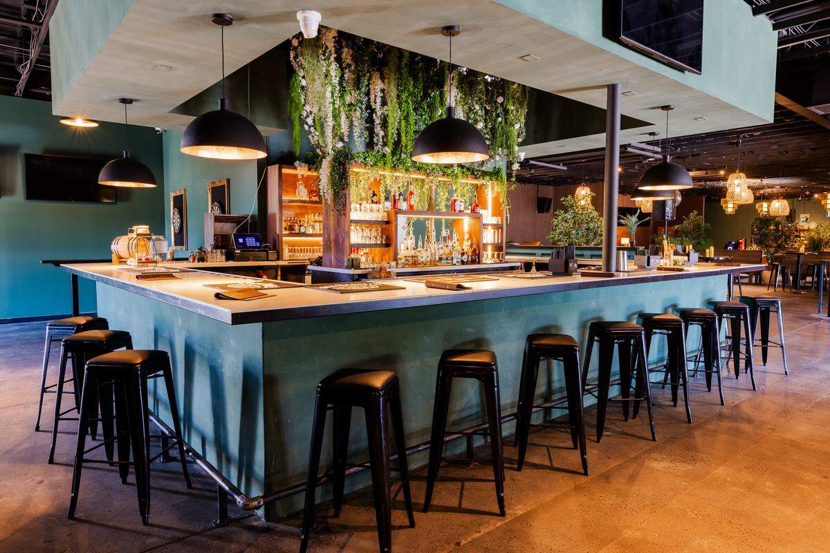 A large, square island bar is surrounded by black metal barstools on a polished concrete floor. The bar structure is painted a muted green and features a light-colored countertop under large, black dome pendant lights with gold interiors. In the center, a tiered wooden liquor display is dramatically draped with cascading artificial greenery and white flowers.