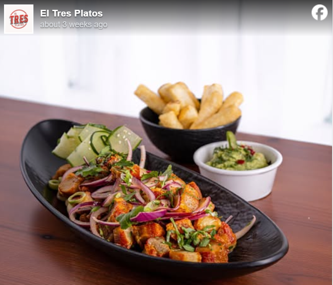A long, black oval dish of crispy pork belly chunks topped with thin slices of red onion, cilantro, and cucumber sits on a wooden table. In the background, a small black bowl holds fried yuca fries, accompanied by a small white ramekin of green salsa or guacamole. The image is a social media post from El Tres Platos, presented with a bright, clean aesthetic.