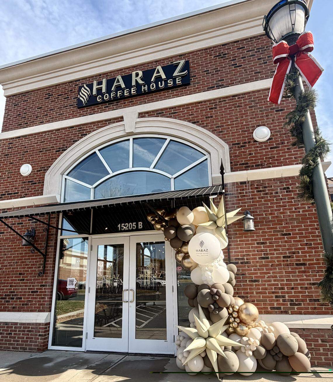 The exterior of the Haraz Coffee House is visible, featuring a brick building facade with a large arched window above the double glass doors. A curved balloon garland in shades of brown, white, and gold decorates the entrance, and a light pole with a red bow stands to the right.