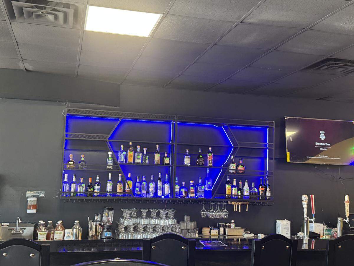 The restaurant’s bar area is shown with a dark counter and a striking, backlit liquor display. Bottles are arranged on shelves accented by bright blue LED lighting against a dark wall, with a TV screen visible on the right.