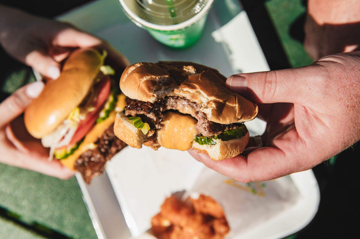 An overhead, close-up shot focuses on two hands holding up burgers above a white serving tray. The burger on the right has a large bite taken out of it, revealing a thick, melted core of yellow cheese oozing from the center of the beef patty. The background is softly blurred, showing a hint of a green cup and a side of golden fries.