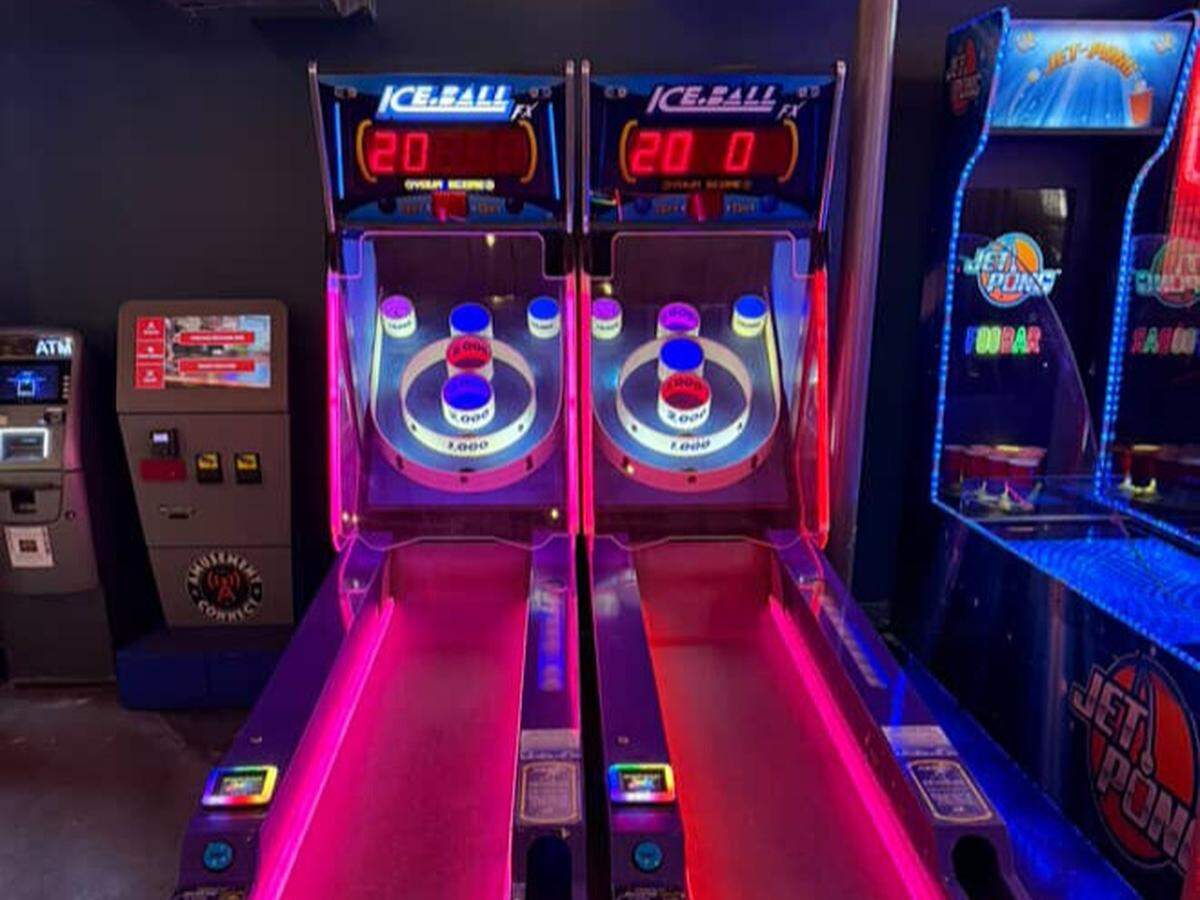 A pair of Ice Ball Skee-Ball arcade machines glows with vibrant pink and blue neon lights in a dark area of the establishment. The classic alley-roller games are ready for players, standing next to an ATM and other arcade cabinets.