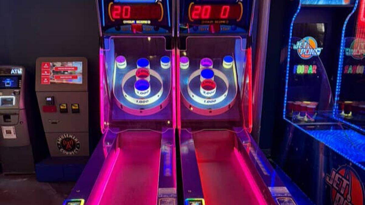 A pair of Ice Ball Skee-Ball arcade machines glows with vibrant pink and blue neon lights in a dark area of the establishment. The classic alley-roller games are ready for players, standing next to an ATM and other arcade cabinets.