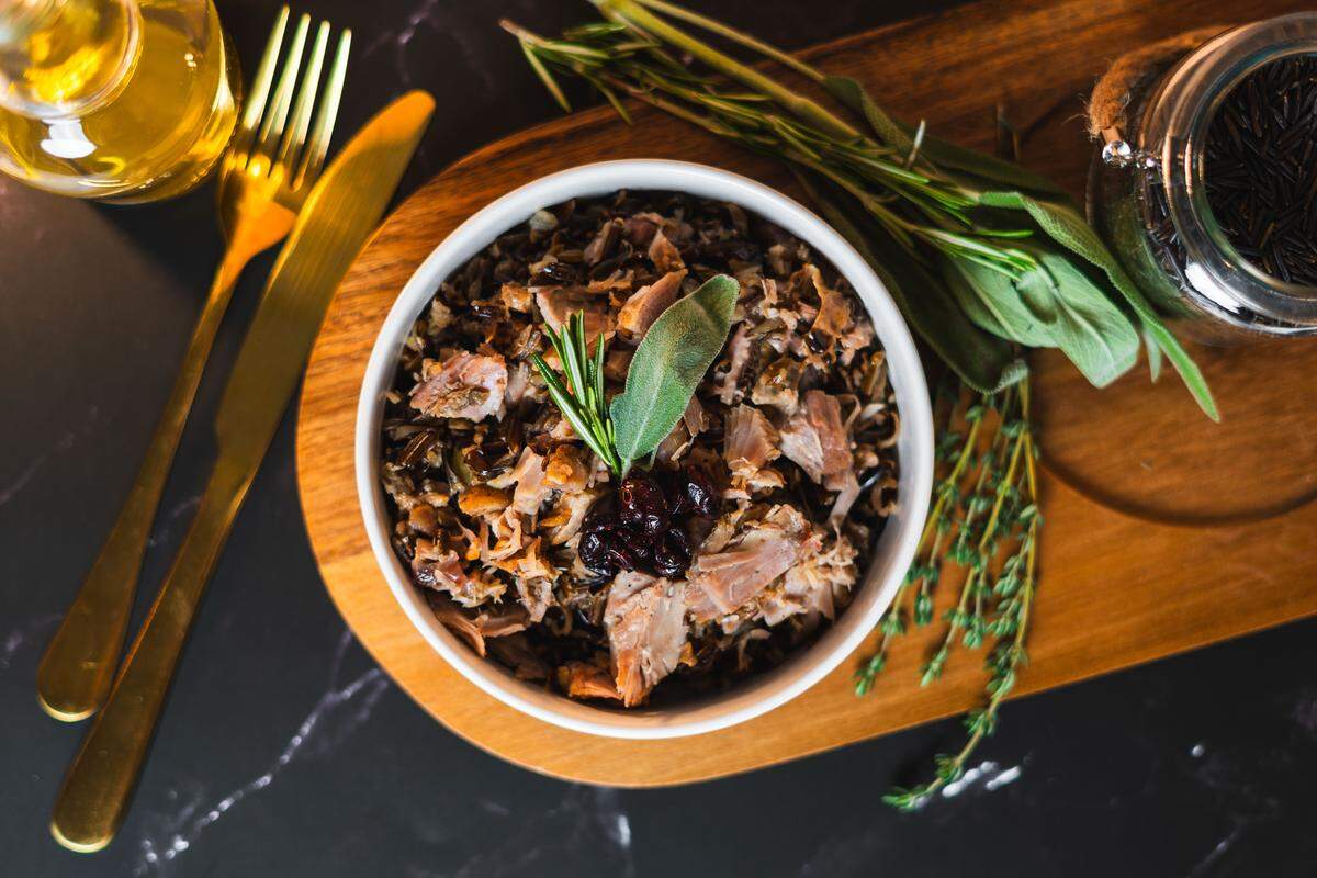 A directly overhead view displays a white bowl of shredded turkey and wild rice resting centrally on a wooden board. The dish is garnished with a sage leaf, rosemary, and dried cranberries, flanked by gold utensils on the left and fresh herbs on the right. A glass jar filled with uncooked wild rice sits in the top right corner against the black marbled background.