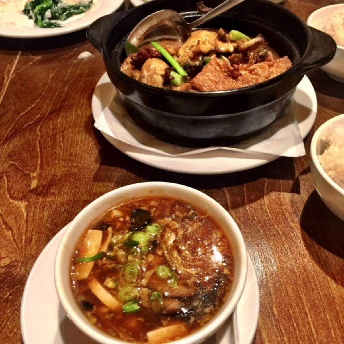 A close-up of a Chinese meal on a dark wood table. In the foreground is a white bowl of hot and sour soup with tofu and scallions. Behind it, a black clay pot contains a savory stew with meat, tofu puffs, and mushrooms, with a ladle resting inside. Side dishes of rice are also visible.