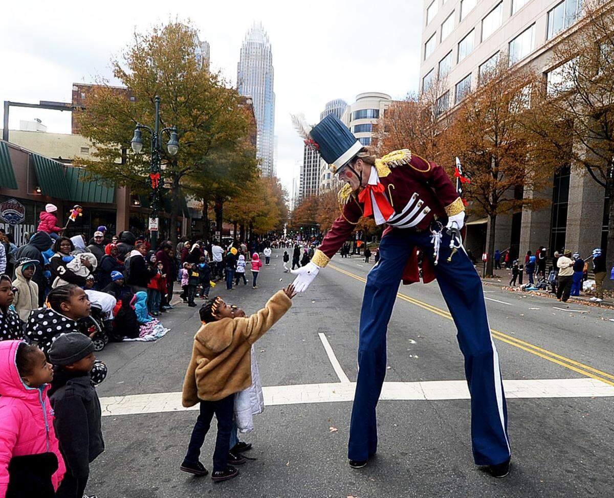 A tall stilt walker dressed in a regal red and blue military-style marching band uniform extends his hand down to shake the hand of a young child looking up at him from the curb. The scene is set on a city street lined with spectators, with modern high-rise buildings and autumn trees visible in the background, likely in Charlotte.