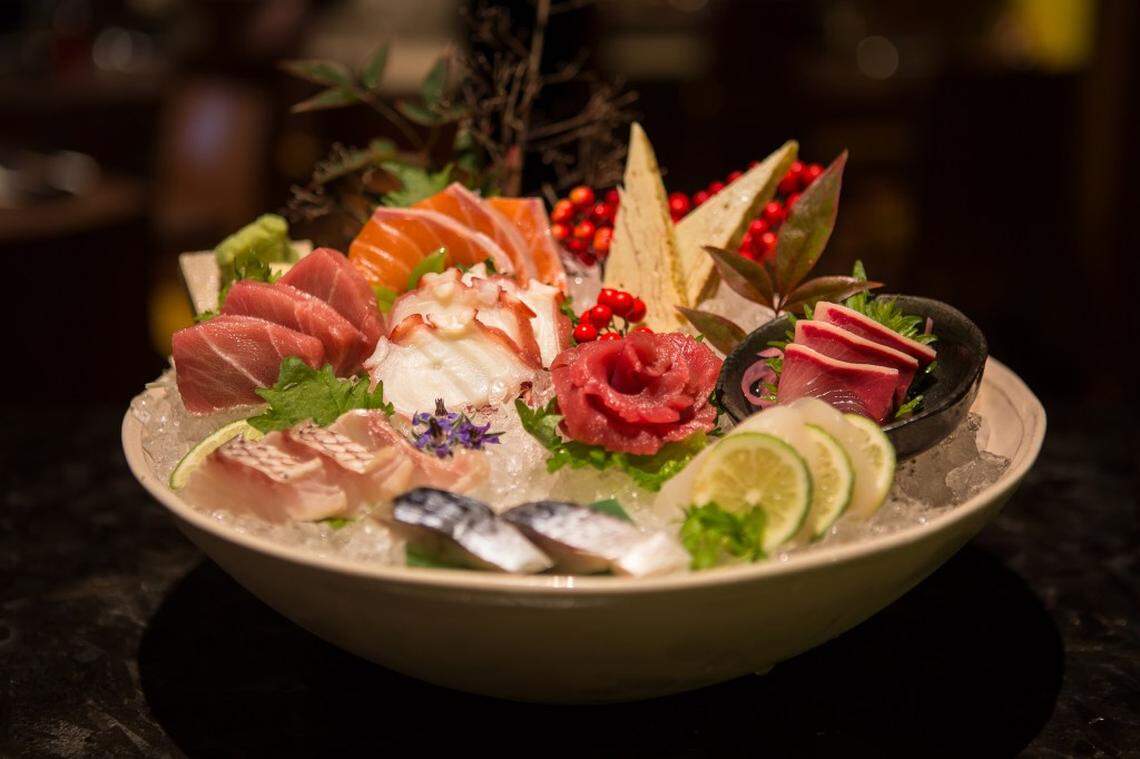 An elaborate sashimi arrangement is presented in a large bowl filled with crushed ice and garnished with red berries and lime slices. The platter features a variety of fresh seafood, including tuna roses, sliced octopus, salmon, and mackerel.