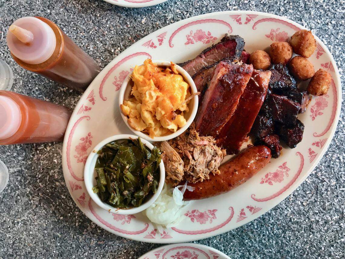 An oval platter of barbecue meats including sliced brisket, ribs, chopped or pulled pork, and a smoked sausage link. The plate also holds two white bowls with sides: macaroni and cheese and collard greens. Small fried hushpuppy-style balls are visible at the top right, and two bottles of barbecue sauce are on the left.
