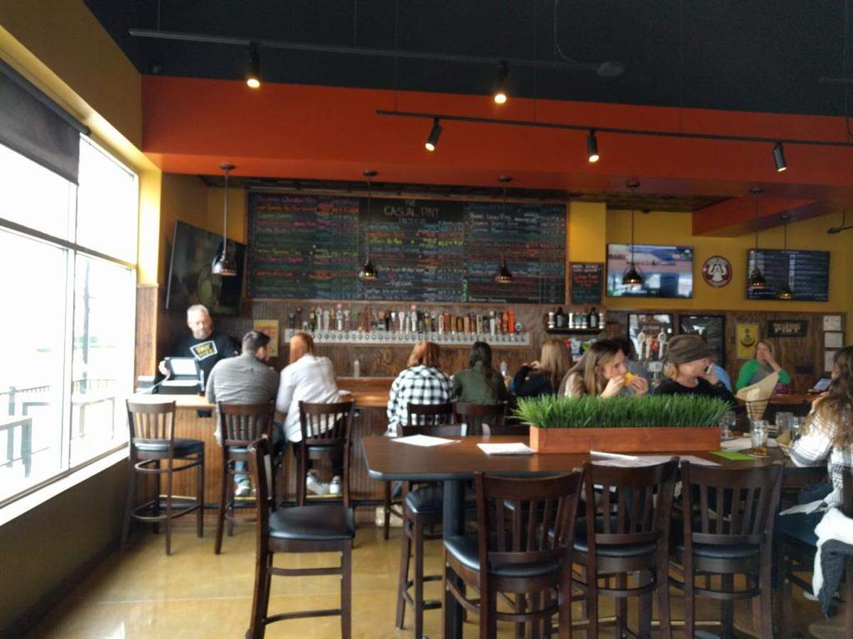 An interior view of The Casual Pint, showing a lively taproom with patrons sitting at a wooden bar and high-top tables. Behind the bar, a large chalkboard menu lists a wide variety of beers in colorful chalk, situated above a long row of silver beer taps.
