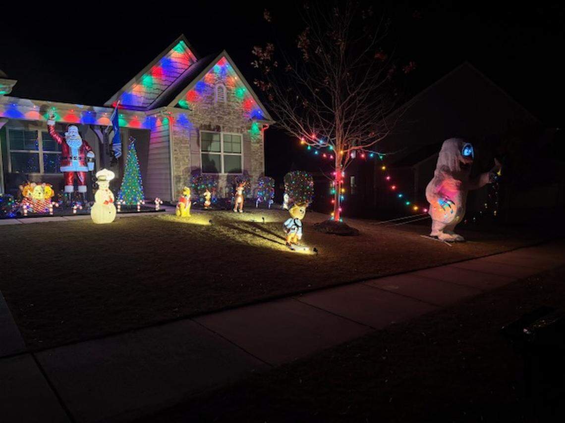 A suburban home at night decorated with a “Rudolph the Red-Nosed Reindeer” theme. In the foreground, an inflatable Bumble (the Abominable Snow Monster) stands on the lawn holding a string of colorful lights that stretch toward a smaller Hermey the Elf figure. To the left, a large, waving Santa Claus figure stands on the porch next to an inflatable Sam the Snowman. The house is accented with colorful spotlights reflecting off the roof, and a small, glowing green Christmas tree sits near the walkway.