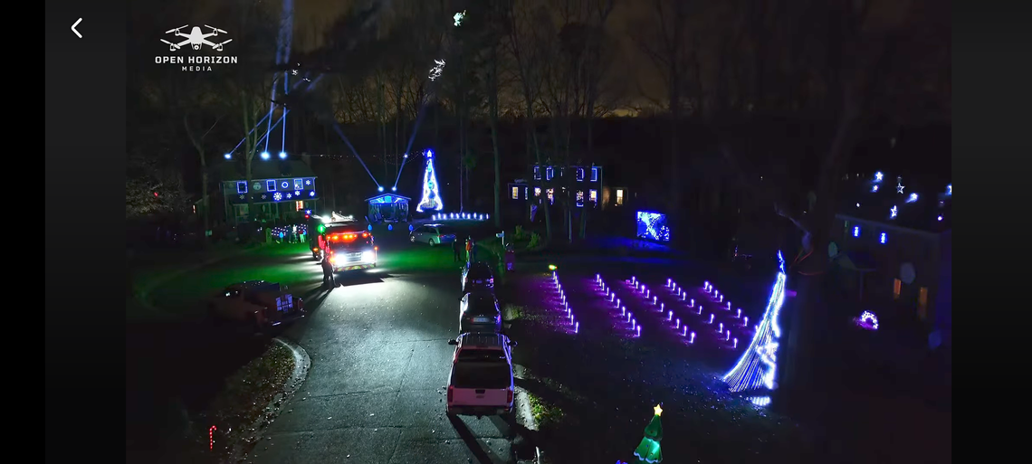An aerial shot shows a neighborhood road at night lined with cars waiting in a queue to view a dramatic Christmas light display. The scene features multiple houses, trees wrapped in blue and white lights, illuminated Christmas tree outlines, and rows of purple lights creating a visual spectacle in the dark.