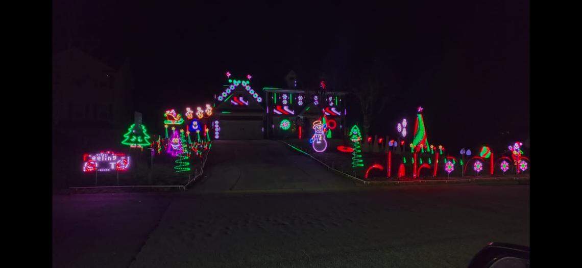 A two-story house is covered in an elaborate, multi-colored Christmas light display featuring neon-style outlines of snowmen, trees, and snowflakes. The vibrant lights line the roof, driveway, and front lawn, creating a glowing spectacle against the dark night sky.