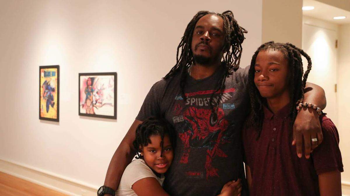 Courtesy of Wolly McNair<br/>Charlotte artist/illustrator Wolly McNair stands in front his artwork on display at the Mint Museum Randolph. Alongside are daughter Malia and son Jayden. McNair’s “Black Hornet” and “of Peace of War” illustrations are part of the Mint’s “Never Abandon Imagination: The Fantastical Art of Tony DiTerlizzi” exhibition, which remains on display through Nov. 3. McNair is one of four local artists featured in the exhibit.