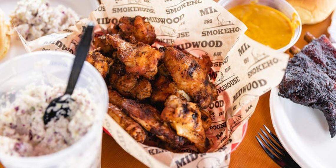 A basket of barbecued chicken wings served in parchment paper, surrounded by side dishes including coleslaw and dipping sauces, with a portion of ribs on a white plate.