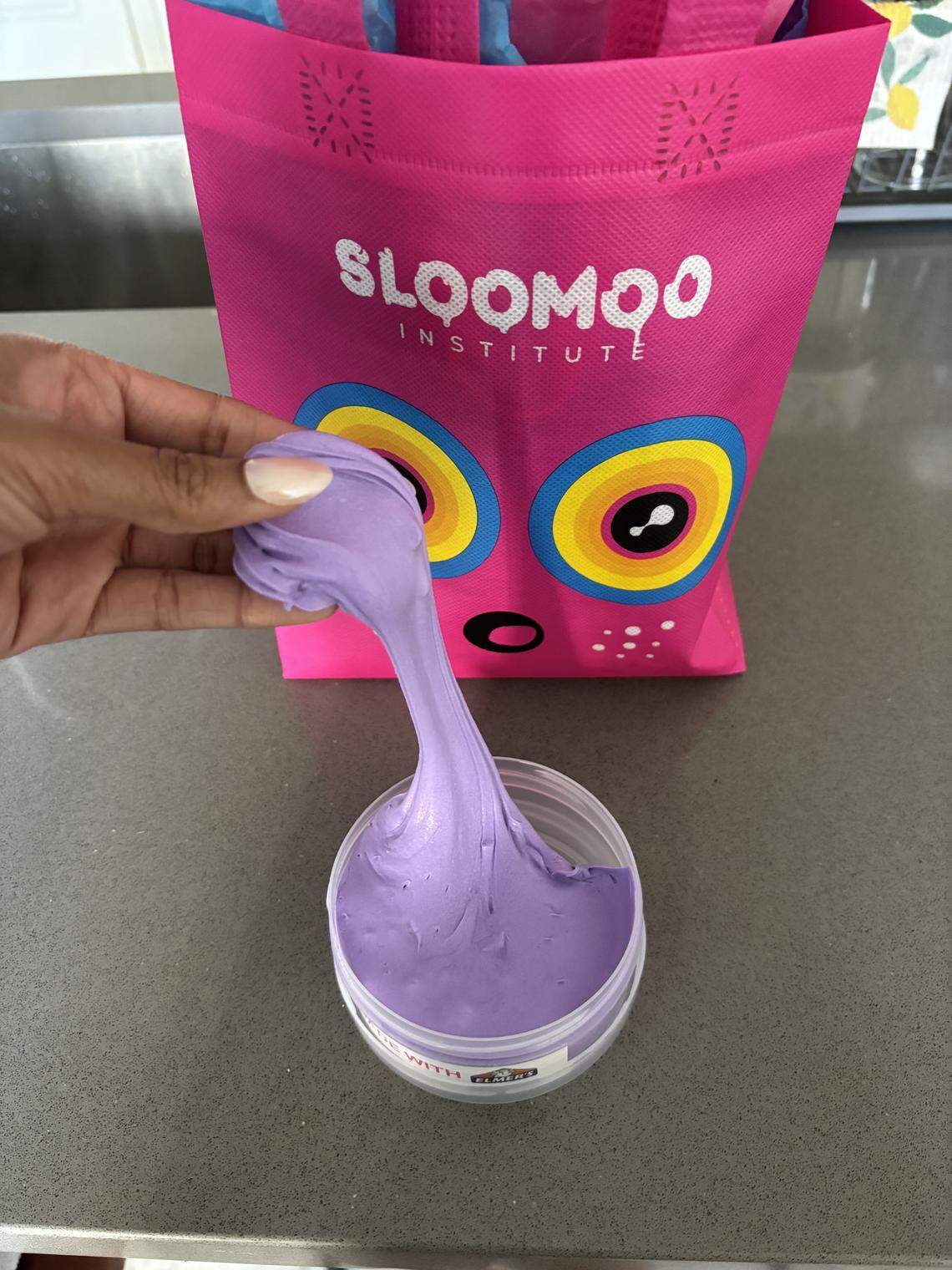 A person pulls a thick, stretchy glob of smooth purple slime from a clear plastic container with their fingers. A pink Sloomoo Institute bag with a whimsical face design stands directly behind the slime on a grey countertop.