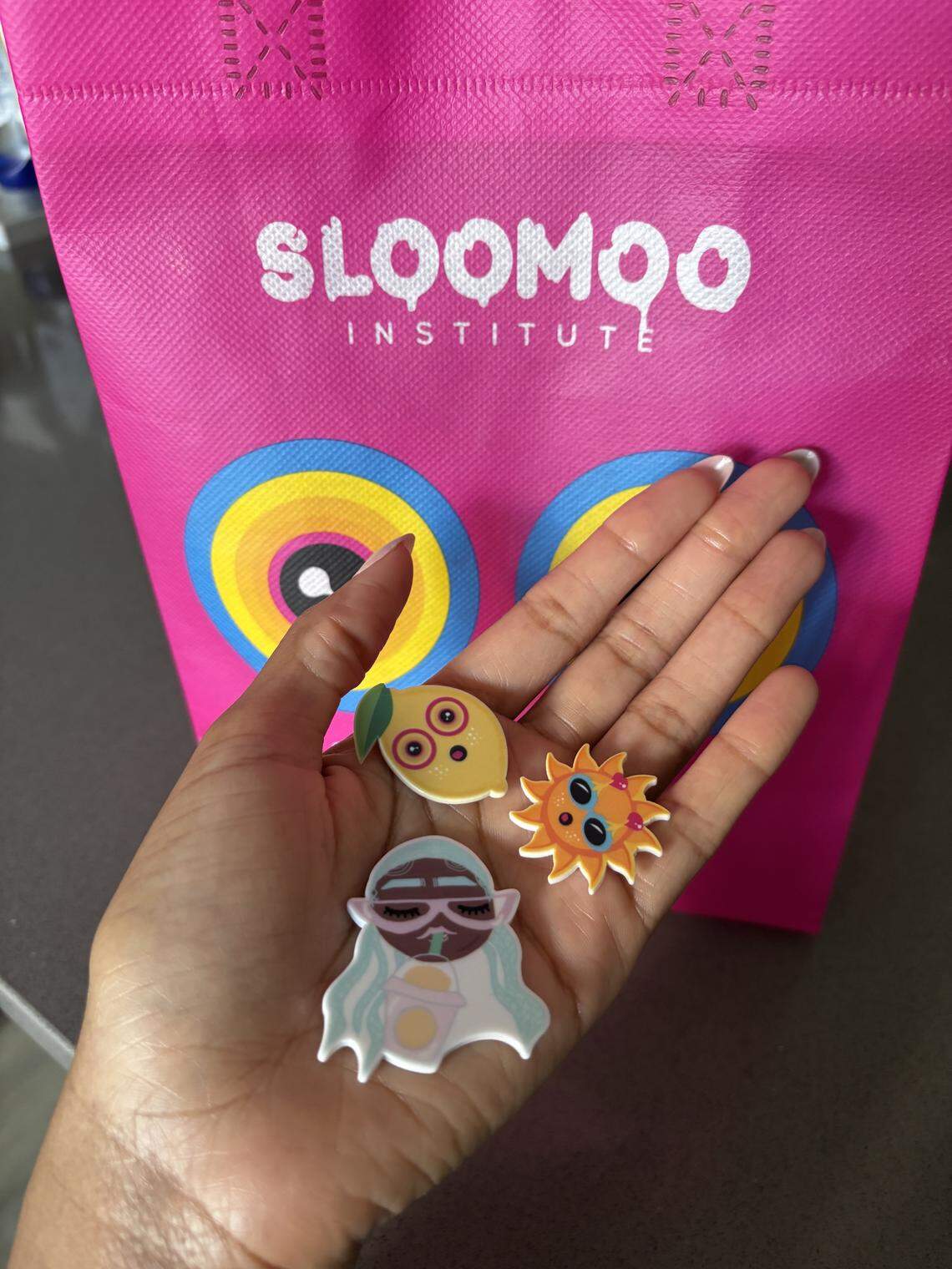 A hand holds three colorful, cartoonish charms—a lemon with eyes, a smiling sun, and a girl with teal hair—in front of a bright pink Sloomoo Institute gift bag. The background features the pink bag’s large, colorful eye logo and the white “Sloomoo Institute” text.