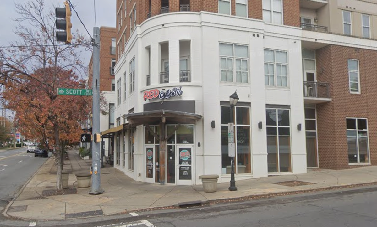 The exterior of “Red @ 28th” is shown on a street corner, featuring a white multi-story facade with large glass windows and a curved entrance under a wooden awning. A green street sign for “Scott Ave” is visible in the foreground near a sidewalk lined with trees and a lamppost.