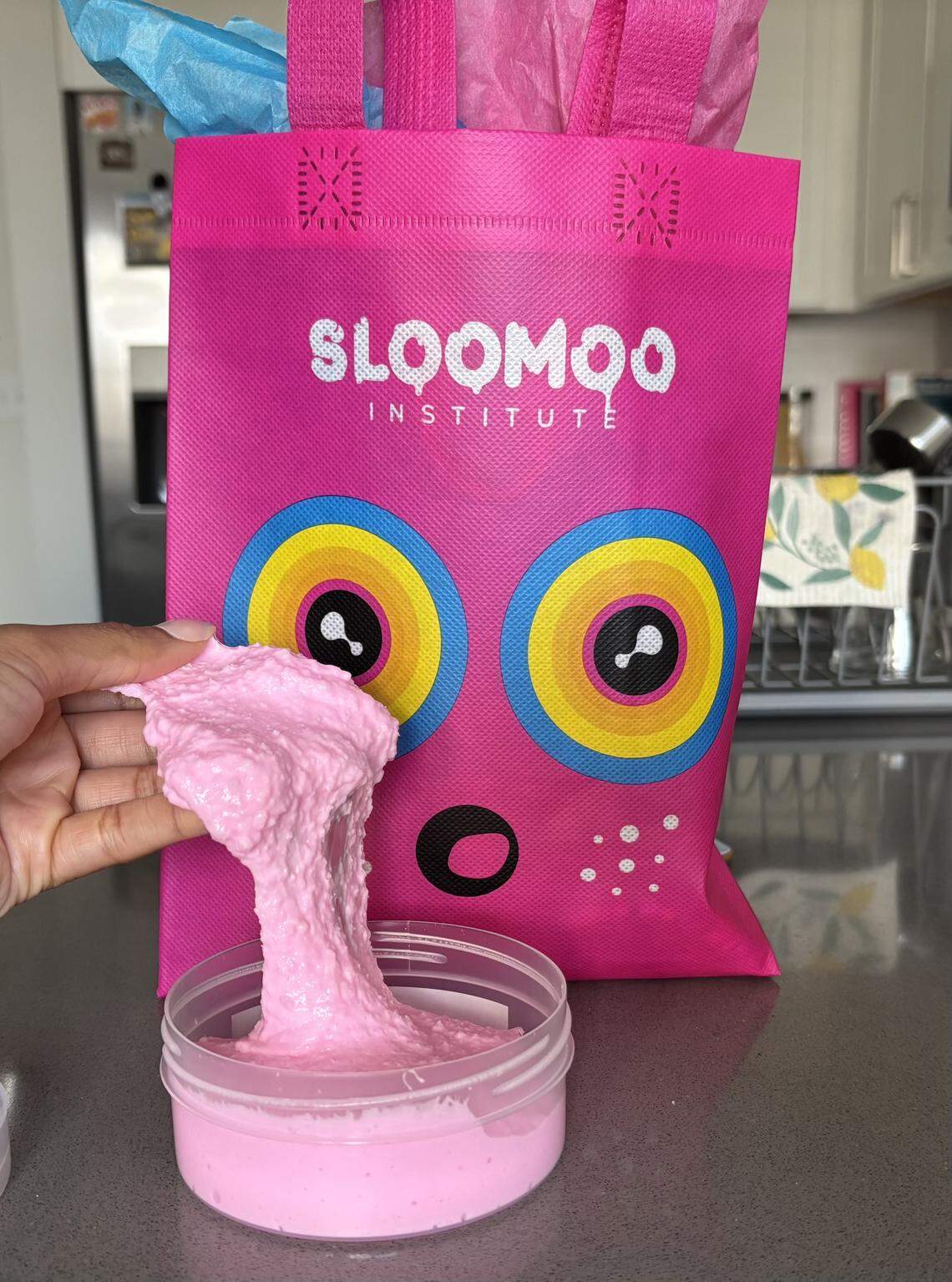 A hand stretches a textured, bubblegum-pink slime upward from its container, showing off its thick and bumpy consistency. The pink Sloomoo Institute bag, filled with blue and pink tissue paper, creates a colorful backdrop for the slime demonstration.