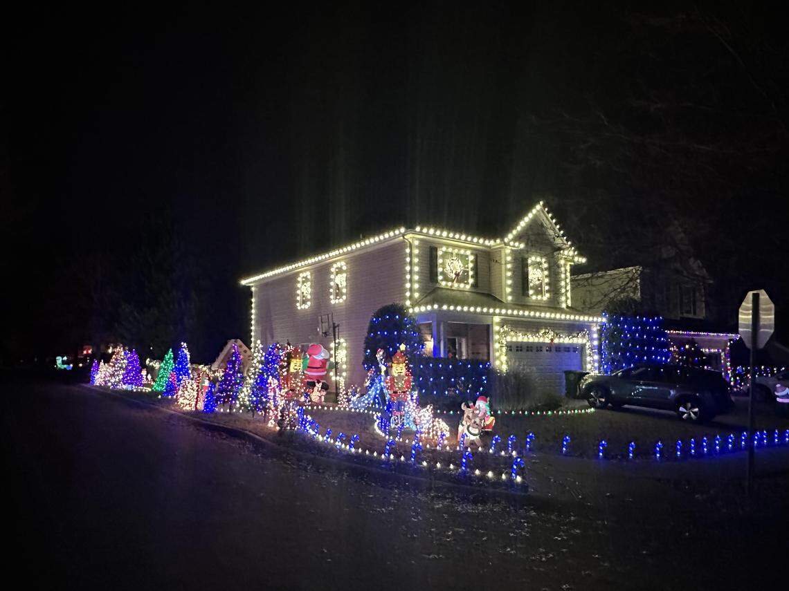A two-story house is densely decorated with bright Christmas lights at night. The roofline and windows are outlined in warm white lights, while the lawn features numerous colorful light-up trees, an inflatable Santa Claus, and nutcrackers. A line of blue lights borders the driveway and sidewalk.