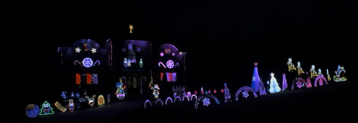 A nighttime photo of a house covered in an elaborate, synchronized display of colorful LED Christmas lights. The display includes arched lights, various cartoon character figures, stars, snowflakes, candy canes, and several Christmas trees glowing in pink, purple, and white light.