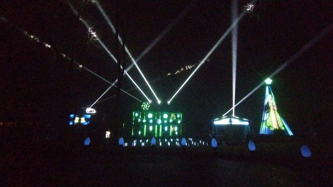 This nighttime photo captures a section of a large, high-tech light display, dominated by powerful white spotlights shining up into the sky. The display includes a central, large structure lit with bright green and blue snowflake projections, flanked by a glowing, conical blue and green Christmas tree outline.
