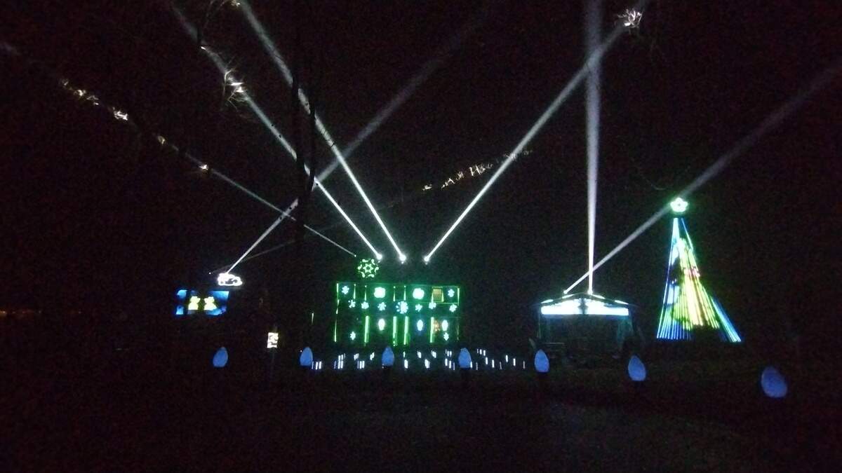 This nighttime photo captures a section of a large, high-tech light display, dominated by powerful white spotlights shining up into the sky. The display includes a central, large structure lit with bright green and blue snowflake projections, flanked by a glowing, conical blue and green Christmas tree outline.
