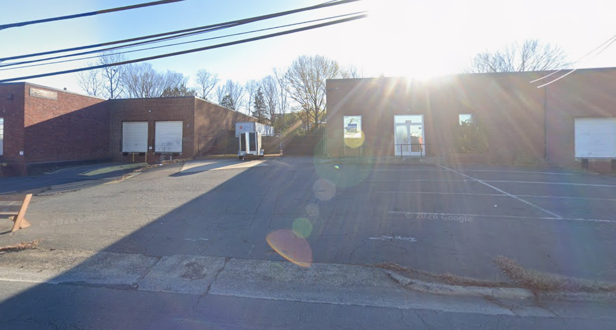 An outdoor daytime view of a single-story brick commercial building with a small parking lot in front, heavily backlit by a bright sun creating a prominent lens flare. To the left, there is a loading dock area with two closed white roll-up garage doors and a small utility trailer parked nearby. Bare trees line the background under a clear sky, and overhead utility lines cross the top left of the frame.