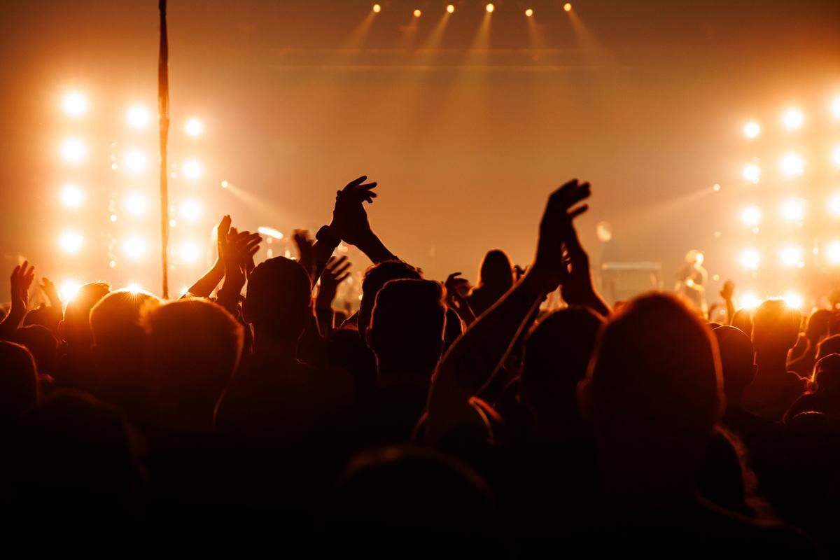 A silhouette of a large crowd is captured from behind as they watch a concert performance under warm, bright stage lights. Many audience members have their hands raised, clapping and cheering in the glowing atmosphere. The stage in the background features multiple vertical light panels casting an intense orange and golden hue over the entire venue.
