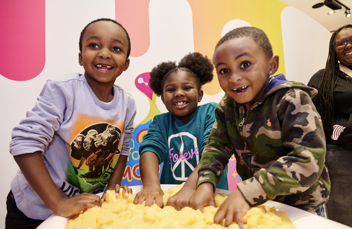 Three joyful children lean over a large container, their hands submerged in a thick, vibrant yellow substance. They are smiling broadly against a colorful, modern backdrop, clearly enjoying a hands-on sensory activity.