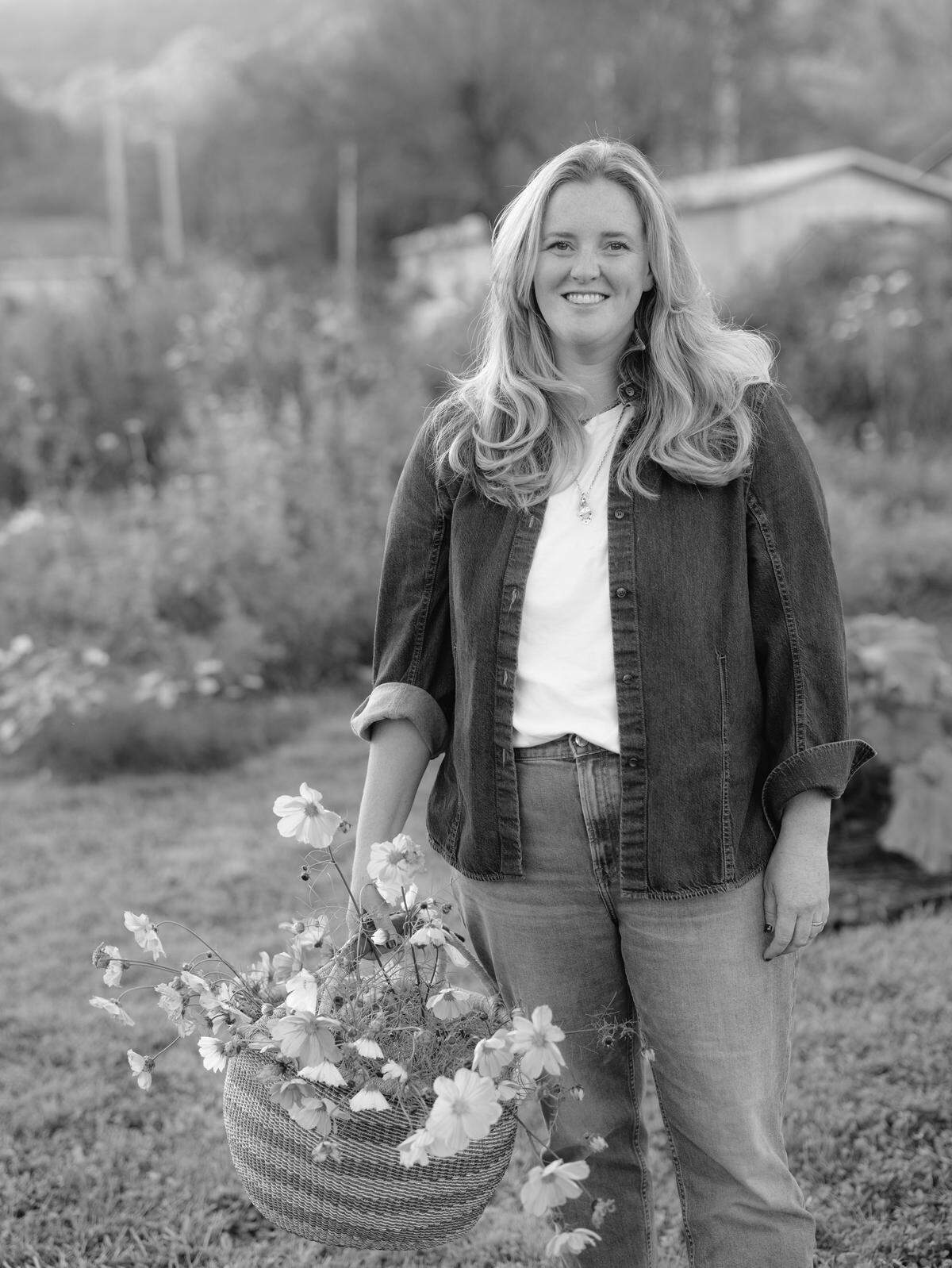 A black and white portrait shows a smiling woman standing outdoors while holding a large woven basket filled with wildflowers. She is wearing a denim button-down shirt over a white tee and jeans, with a soft-focus garden and trees in the background.