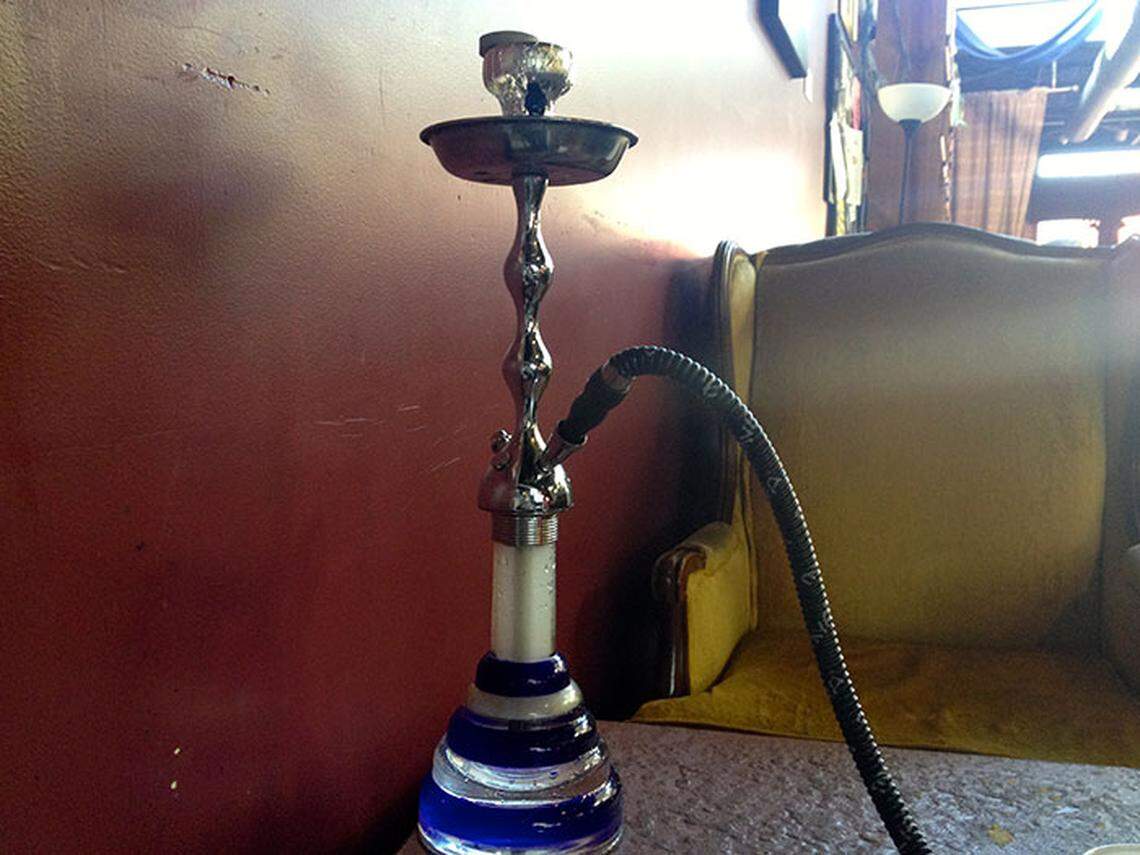A silver and blue glass hookah sits on a textured table against a deep red wall. In the background, a plush yellow armchair and a warm floor lamp create a cozy lounge atmosphere.