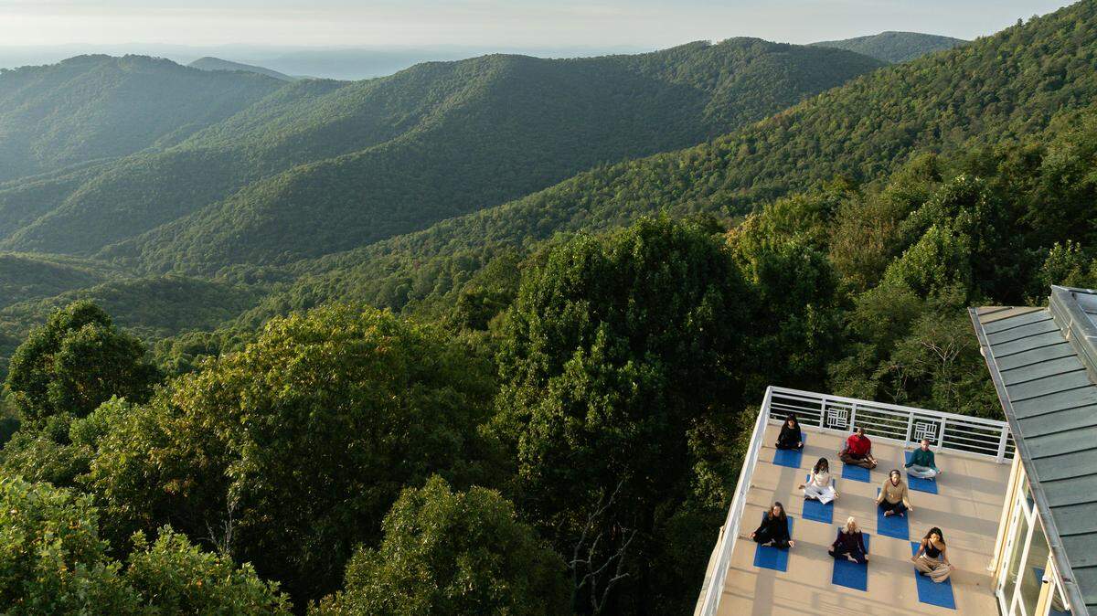 An aerial view shows a group of about nine people sitting on yoga mats on a terrace balcony overlooking a vast expanse of lush, green, rolling mountains. The people are facing away from the camera, meditating or resting in the stillness of the beautiful, hazy mountain landscape under a light blue sky.