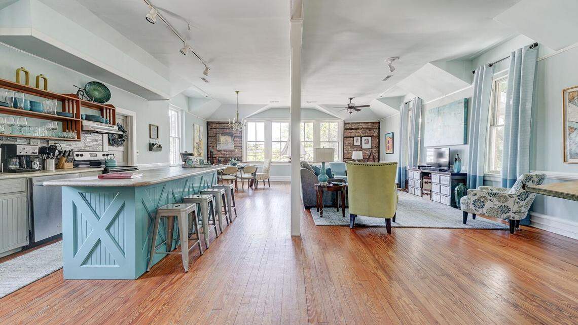 This wide-angle shot shows the open-concept transition from a teal kitchen island to a spacious living and dining area. Track lighting runs along the ceiling, illuminating the long wooden floorboards that lead toward a sunlit dining nook and a cozy sitting area. The layout emphasizes the historic building’s airy feel and the cohesive “coastal industrial” decor.