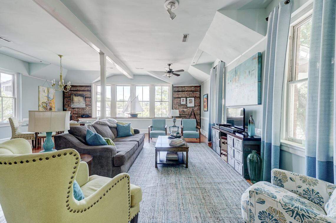 An expansive living room features light blue walls, original hardwood floors, and exposed brick accents framing large windows. A dark grey sofa and various accent chairs in seafoam green and patterned blue are arranged around a central wooden coffee table. Natural light pours in, highlighting a large sailboat model on the windowsill and soft blue drapes.