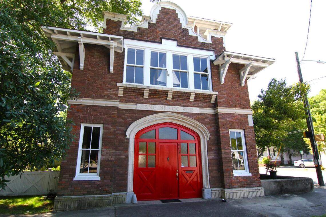 The front of the historic “Engine Co. No. 2” firehouse is a two-story red brick building featuring a striking, arched bright red double door. White architectural trim and decorative brackets support the roofline, while a row of four large windows on the second floor looks out over the street. The building is flanked by lush green trees and a clear, sunny sky.