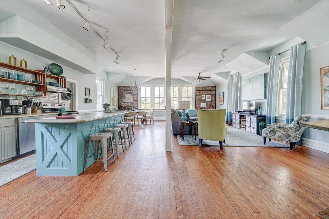 This wide-angle shot shows the open-concept transition from a teal kitchen island to a spacious living and dining area. Track lighting runs along the ceiling, illuminating the long wooden floorboards that lead toward a sunlit dining nook and a cozy sitting area. The layout emphasizes the historic building’s airy feel and the cohesive “coastal industrial” decor.
