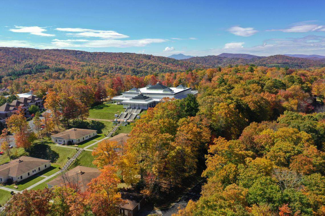 This is an aerial shot capturing a large, distinctive white building surrounded by a densely forested landscape vibrant with the red, orange, and yellow foliage of peak autumn. Several smaller residential buildings are visible in the foreground, with the colorful trees and mountains stretching out under a bright blue, partially cloudy sky.