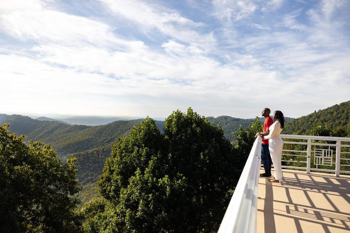 A side profile view of a Black man and woman standing at a white railing, gazing out at the extensive mountain vista and blue sky. The couple is standing on a high-elevation observation deck overlooking a sea of green treetops and distant rolling hills on a clear, sunny day.