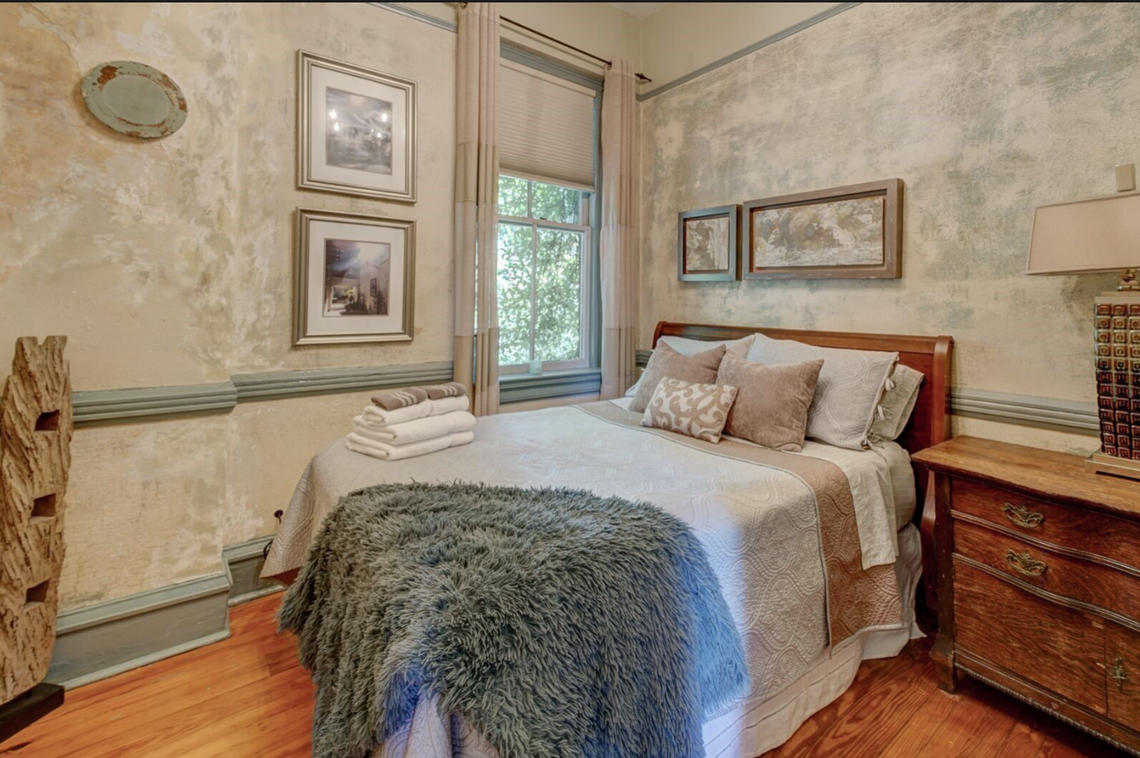 A cozy bedroom features weathered, textured walls in shades of beige and blue that give the space a vintage, rustic feel. The bed is dressed in neutral linens with a shaggy grey faux-fur throw and is flanked by a classic wooden dresser with an ornate lamp. Soft light filters through a single window, illuminating framed artwork and a stack of fresh white towels on the bed.